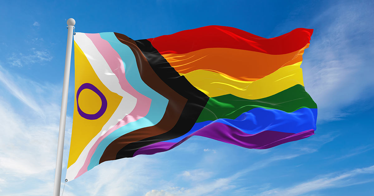 Image of the progress pride flag, including the original rainbow colors as well as a yellow triangle with a purple circle for the intersex pride flag, brown and black stripes recognizing people of color in the LGBTQ+ community, and blue, pink, and white stripes representing the Trans Pride flag.