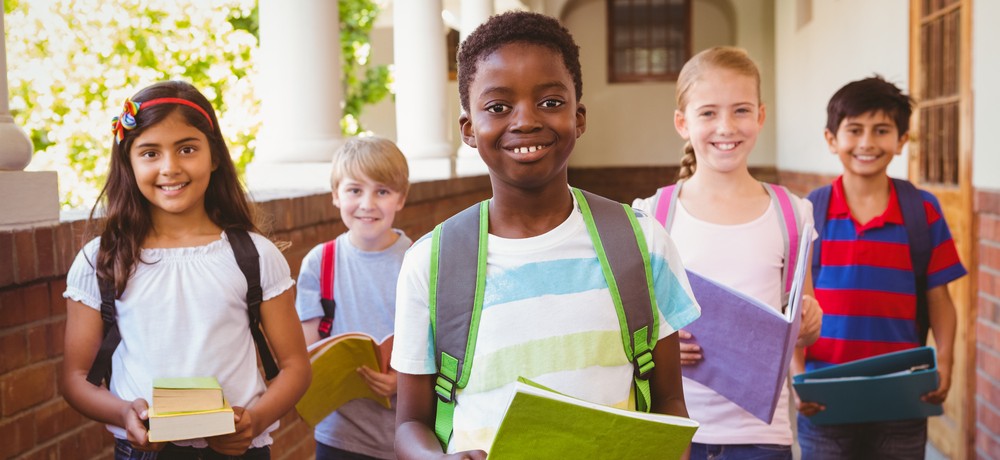 A group of children are standing together outside a school. They are wearing backpacks, holding books and binders, and smiling at the camera.