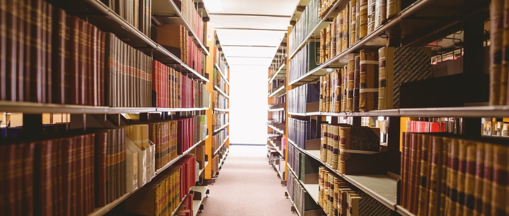 An picture of a narrow aisle between shelves of scholarly books in a library.