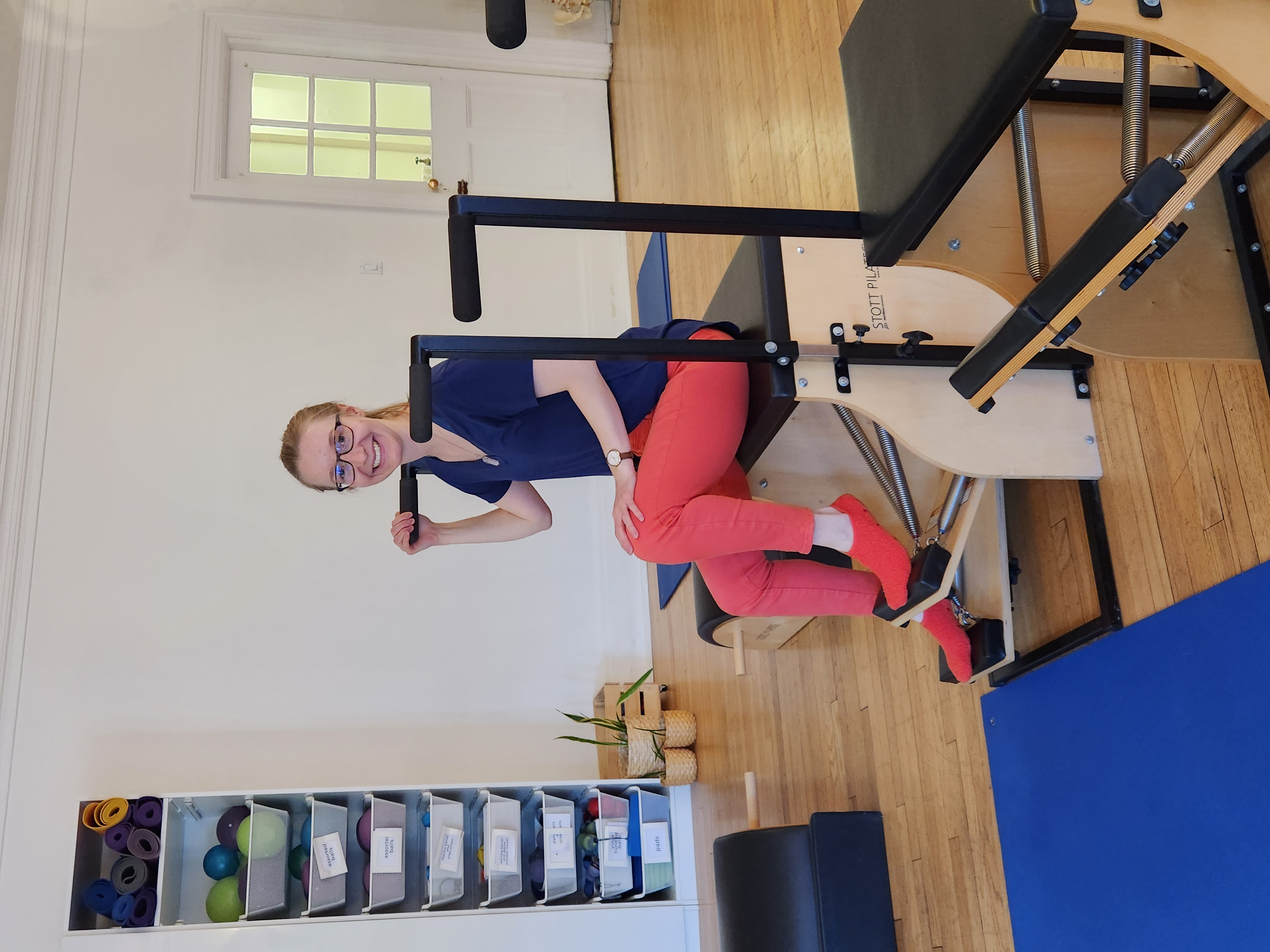 Emily is sitting on the PIlates chair smiling. She is wearing a blue shirt and red leggings and socks.